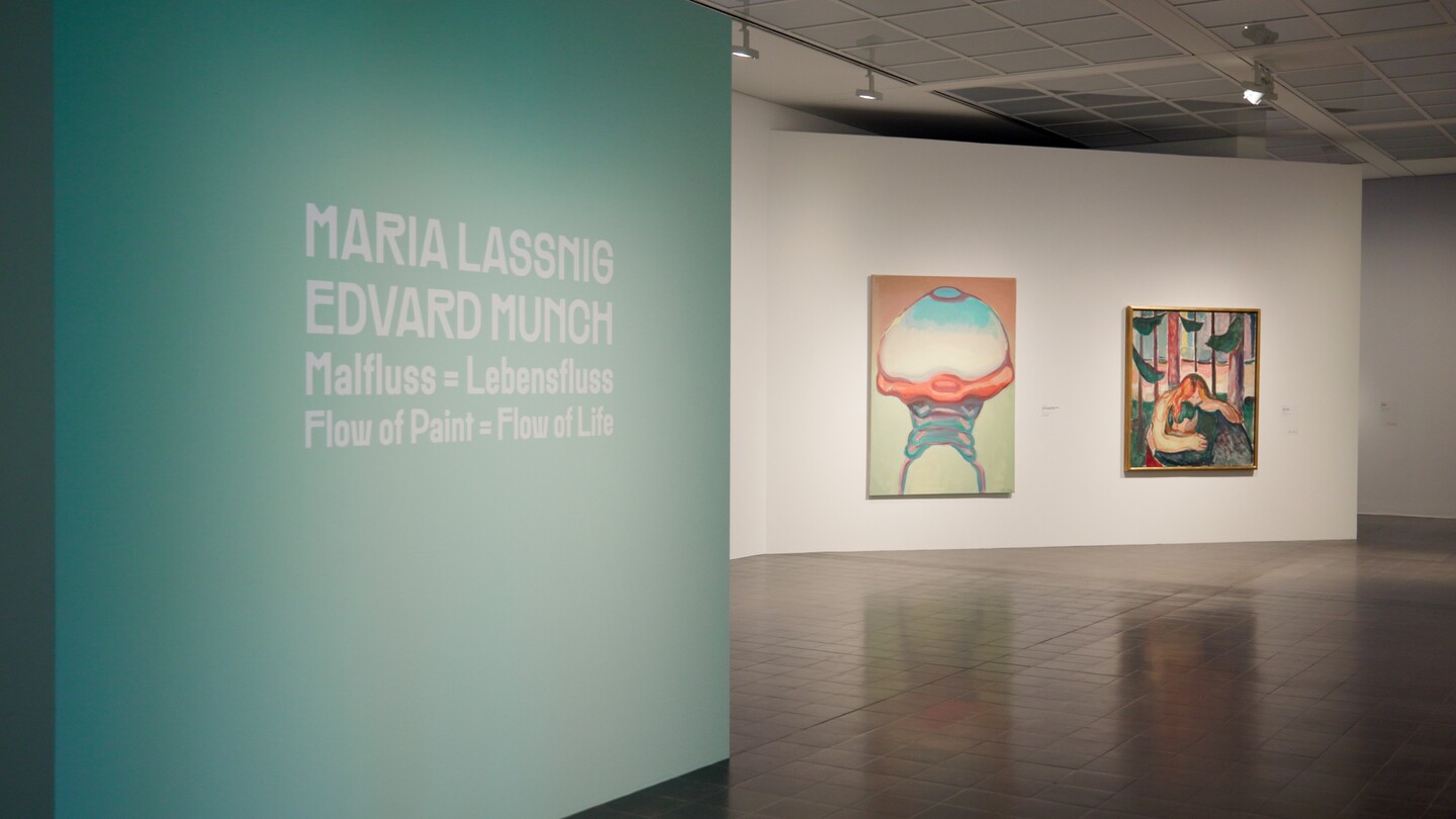 Galerieraum mit türkiser Wand, Text und zwei gerahmten Kunstwerken an weißer Wand