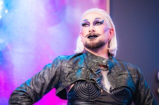 Drag Queen mit rosa Haaren und Make-Up in Lederjacke vor buntem Hintergrund