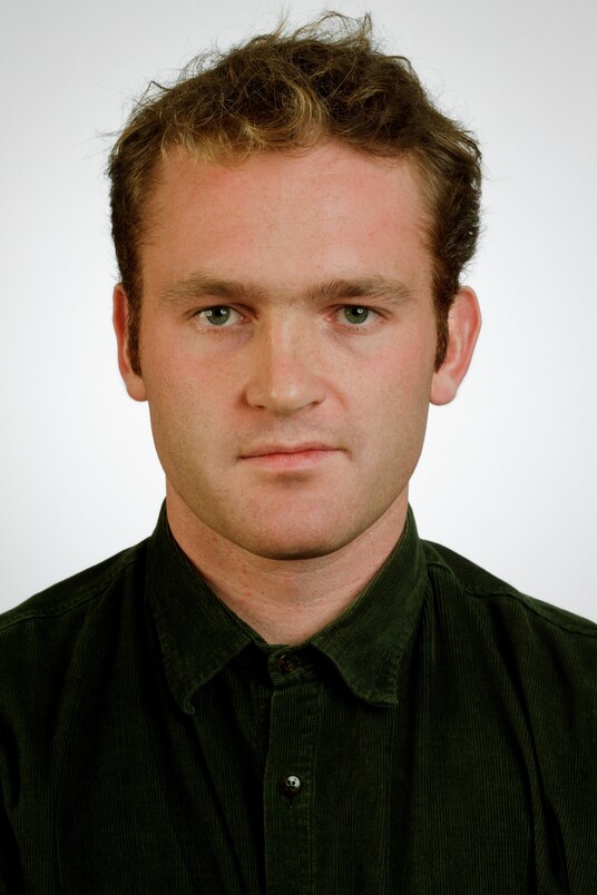 Portraitfoto eines jungen Mannes mit braunem Haar in schwarzem Poloshirt vor weißem Hintergrund